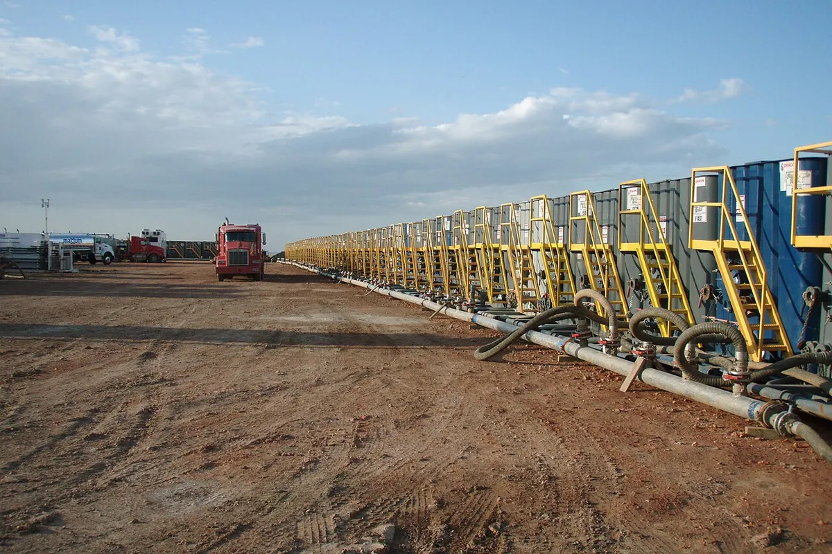 Row of frac water tanks with hoses on a red-dirt location, with trucks in the distance